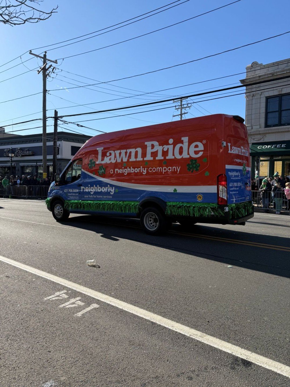 Lawn Pride van decorated with shamrocks and green fringe driving in the St. Patrick’s Day Parade in Bay Shore, with crowds gathered along the street.
