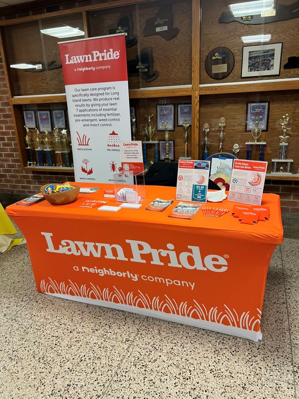 Lawn Pride booth at Hauppauge High School Fair with branded orange table cover, informational brochures, and display banner promoting lawn care services.