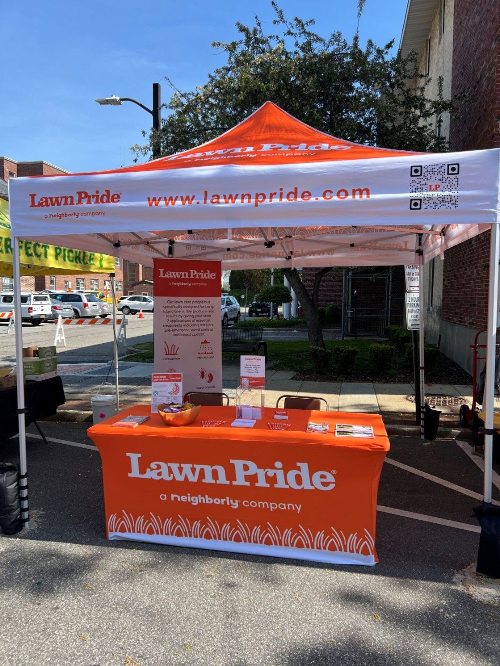 Lawn Pride booth at Oyster Bay Day 2025 with orange canopy and table cover, brochures, and promotional materials promoting lawn care services.