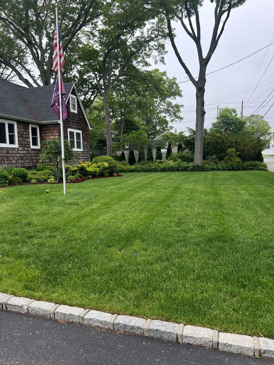 Green, well-manicured front lawn bordered by a stone curb, with a flagpole and landscaped flower beds in front of a shingle-style home on a cloudy day.