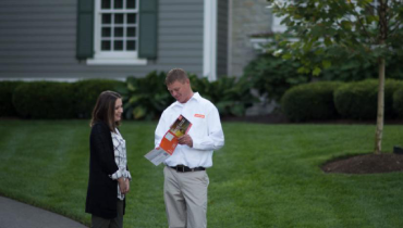 A man shows brochures to a woman outside a house.